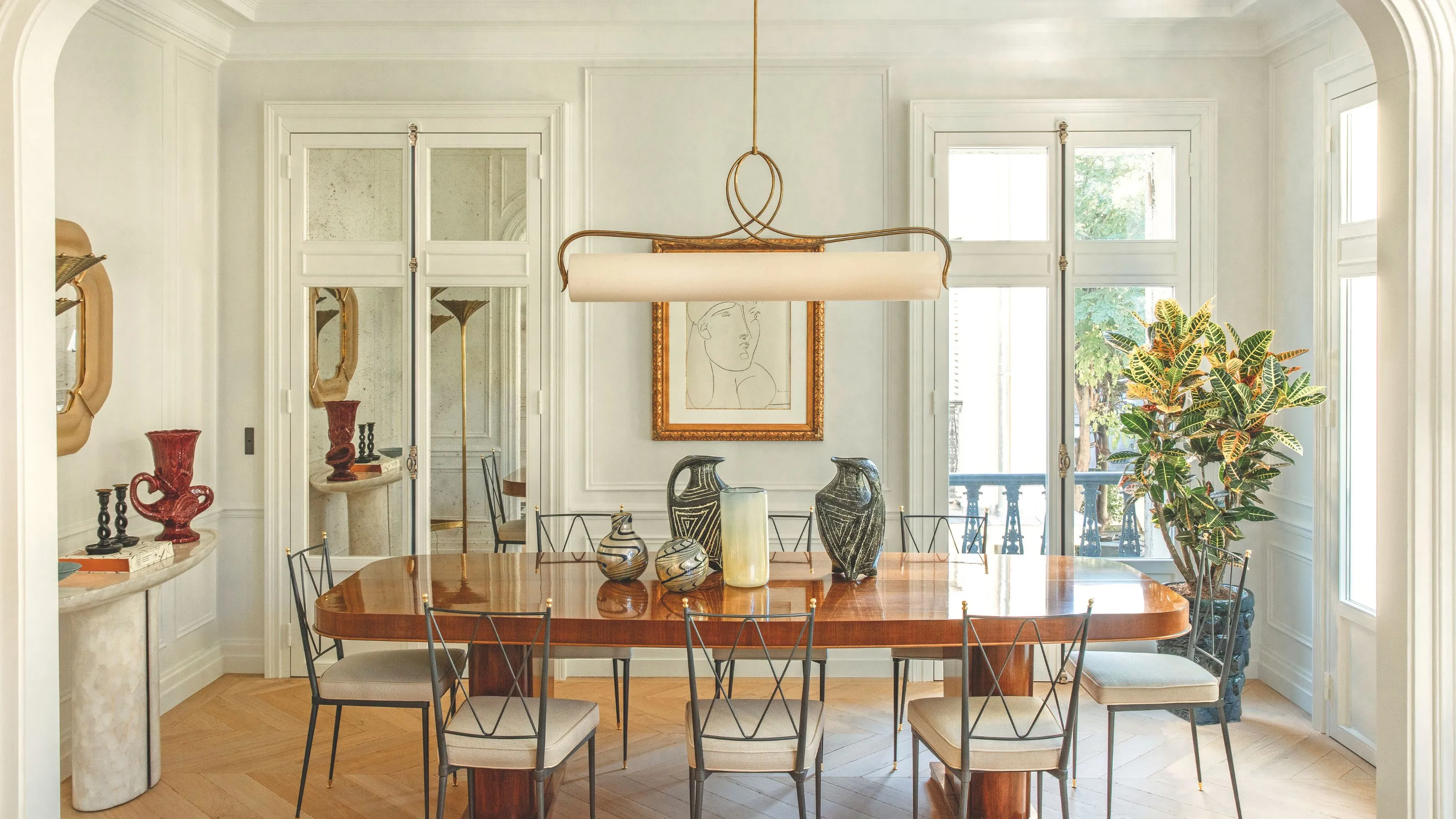 En elegant solbelyst spisestue med et bord i polert tre, slanke svarte metallstoler og en unik horisontal pendellampe. Rommet er dekorert med keramikkvaser og en stor innrammet skisse på panelvegg.' loading='lazy' title=