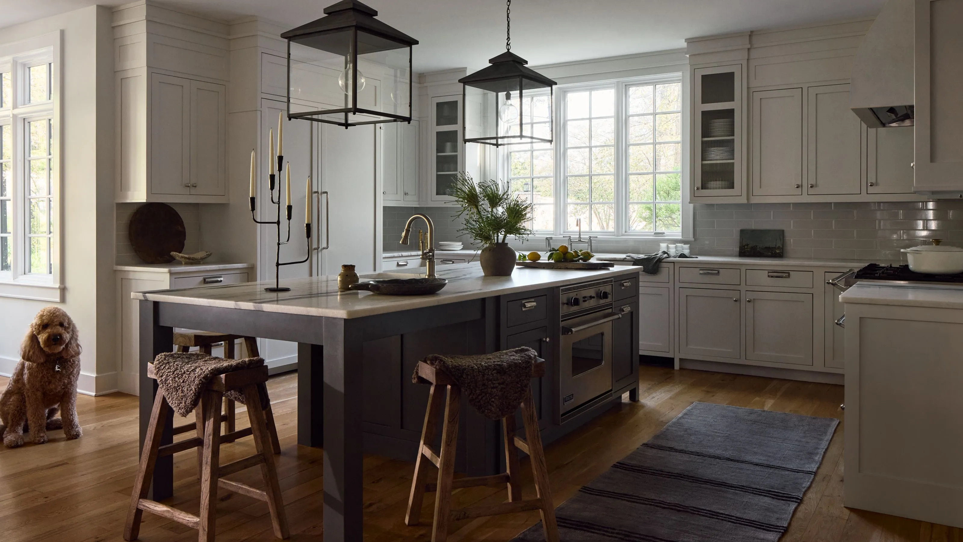Grande cuisine avec armoires murales gris pâle et un îlot gris foncé au centre. Deux suspensions lanternes sont suspendues au-dessus et des outils en bois rustiques permettent de s'asseoir à une extrémité.' loading='lazy' title=