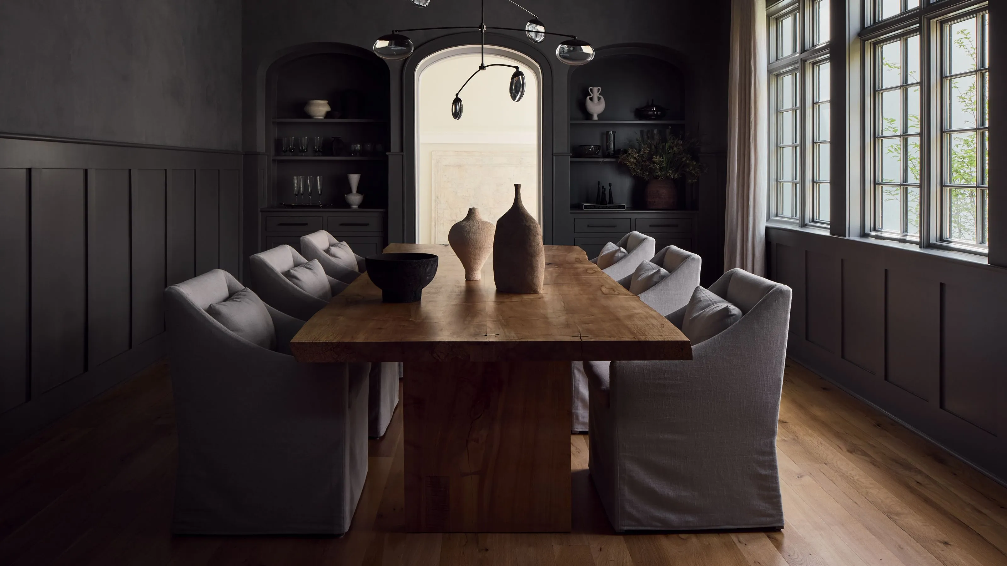 Salle à manger sombre peinte dans un gris anthracite maussade avec des lambris de la même couleur à mi-hauteur des murs. Il y a une grande table en bois au milieu avec des chaises de salle à manger rembourrées grises du même gris et une grande fenêtre remplit la pièce de lumière.' loading='lazy' title=