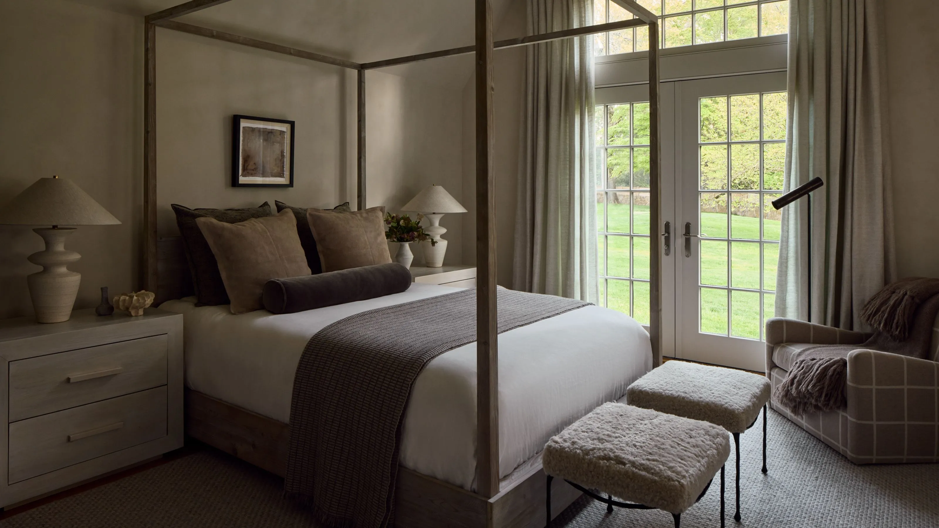 Chambre neutre avec lit à baldaquin et grandes portes-fenêtres. Il y a de longs rideaux autour des fenêtres et le lit est décoré de plusieurs draps neutres.' loading='lazy' title=