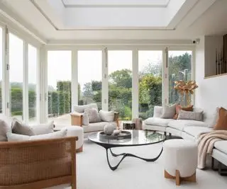 Sala decorada em cores neutras, com móveis em tons naturais de madeira. As janelas do chão ao teto oferecem vista para o jardim, criando uma atmosfera relaxante