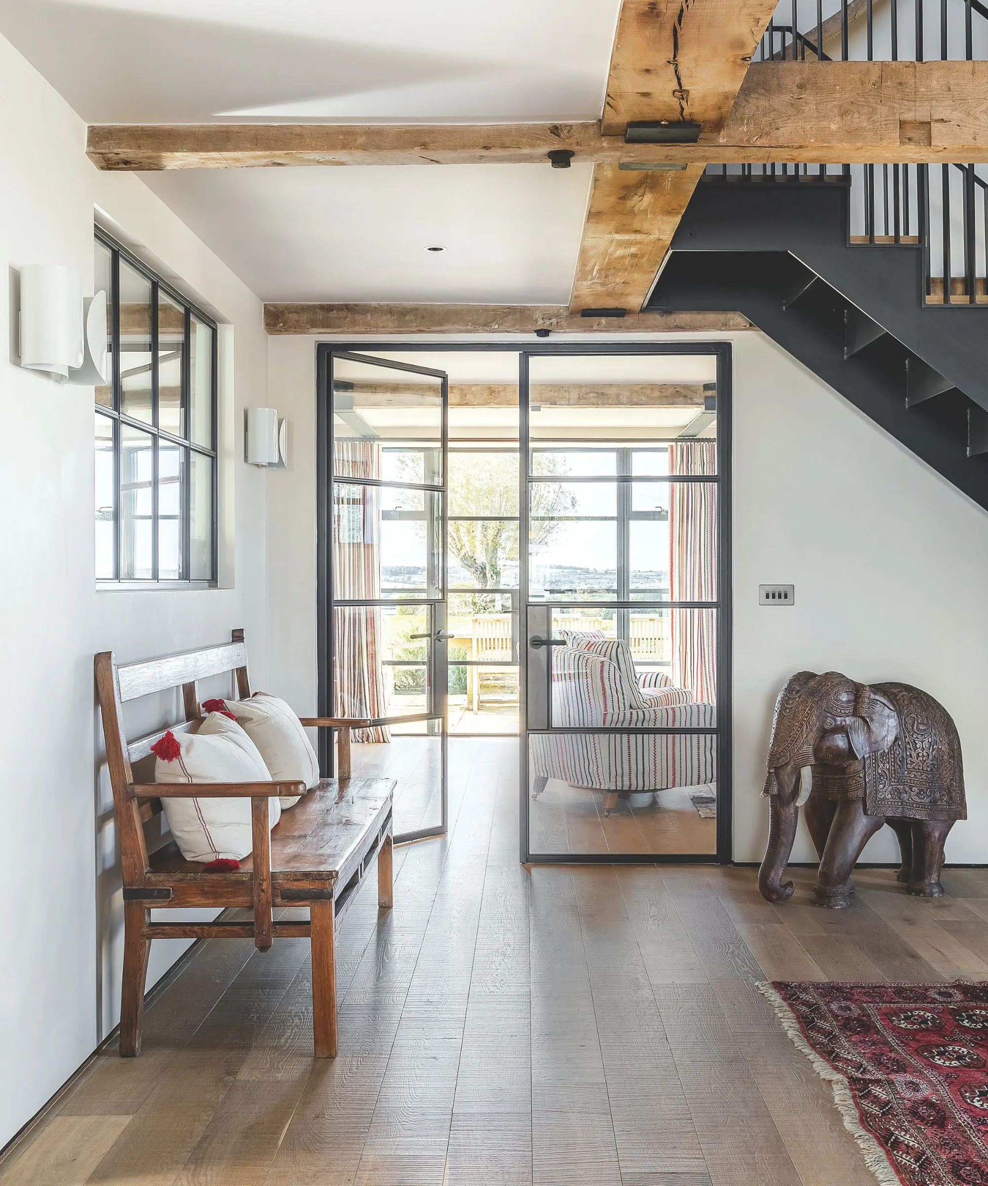 Chodba s drevenými trámami kovové schodisko drevená podlaha s červeným perzským kobercom, starožitná socha slona a drevená lavica' loading='lazy' title=