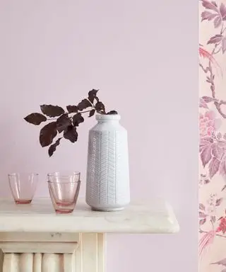 Nærbilde av lilla malt vegg over peisen, floral lilla tapet til høyre, hvit vase med tørkede lilla blomster, små rosa glassglass