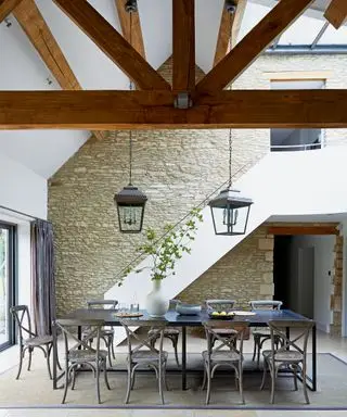 Grande salle à manger avec plafond voûté à double hauteur, poutres en bois sombres, mur en pierre, grande table à manger en bois sombre avec neuf chaises, deux suspensions de style lanterne en métal noir et verre, tapis au sol pour zoner l'espace