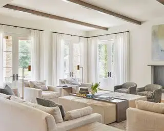 Grand salon avec poutres apparentes en bois sombre, murs peints en blanc, grand coin salon avec canapés et fauteuils crème, deux petits fauteuils gris, grand pouf avec plateau d'objets décoratifs et accessoires