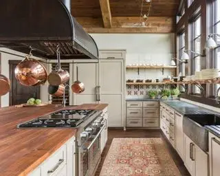 Grande cuisine rustique, armoires grises, îlot de cuisine avec comptoir en bois, support à casseroles au-dessus, étagères décorées d'ustensiles de cuisine et de verrerie, tapis sur sol en pierre, plafond avec poutres apparentes en bois