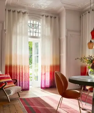 Stor traditionel spisestue med farverigt, moderne design. Ombre pink, orange og hvide gardiner, lyse trægulve, pink stribet tæppe, afrundet metal spisebord med fløjl bille spisebordsstole