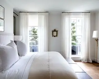 Dobbeltseng i lysegråt soveværelse med trægulv, creme- og hvidt sengetøj og hvide gardiner med gulv til loft-vinduer.