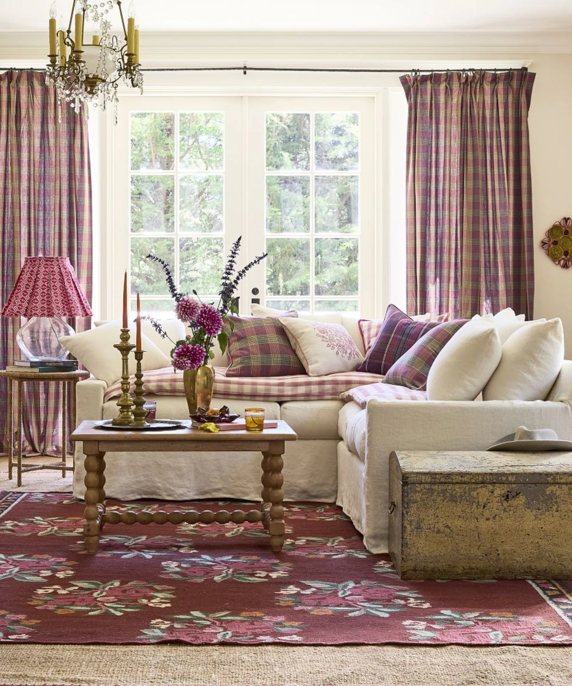 neutralny salon z dużymi francuskimi drzwiami i białą narożną kanapą ozdobioną różowymi poduszkami w kratę z dopasowanymi zasłonami, drewnianymi meblami w stylu vintage i kwiatowym dywanikiem tkanym na płasko' loading='lazy' title=