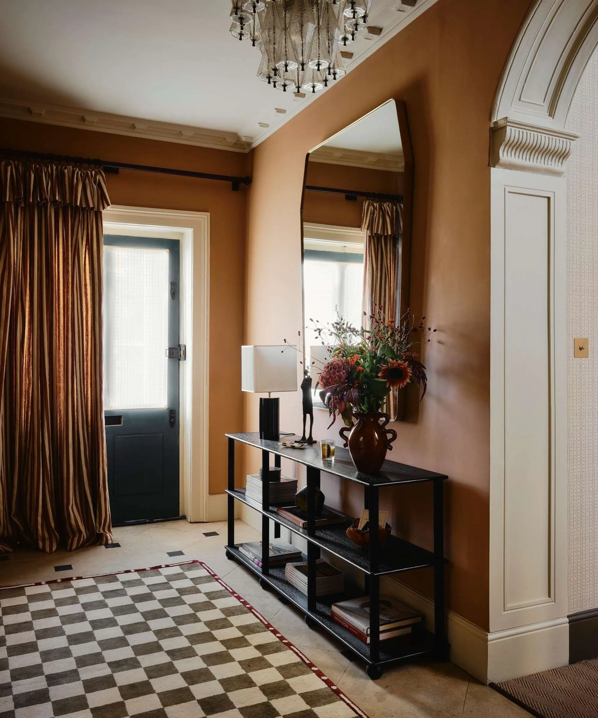 Cálido pasillo ocre con suelo de baldosas, alfombra de tablero de ajedrez, gran espejo y mesa consola, viñeta y una cortina de puerta delantera con volantes a rayas' loading='lazy' title=