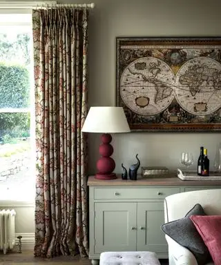 Biblioteca no hall de entrada com piso em madeira, armário em forma de casca de ovo, repousa-pés estofado bege e janela com cortinas com motivos florais