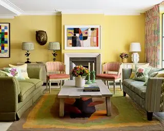 Sala de estar colorida com parede pintada de amarelo, dois sofás verdes, duas cadeiras vermelhas, mesa de centro baixa de madeira, obras de arte coloridas montadas acima da lareira, três luminárias de mesa na mesa console