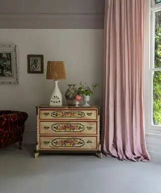 Chambre traditionnelle avec tapis gris, commode en bois peint, décorée avec lampe de table, rideaux roses, grande baie vitrée, murs peints en blanc, élément peint en gris
