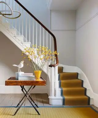 Couloir coloré avec rampe d'escalier et tapis jaunes, console en bois avec fleurs jaunes, livres et ornements, murs peints en blanc, parquet, luminaire arrondi contemporain