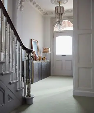 Couloir avec tapis de style velours bleu clair, murs peints en blanc, élégant lustre en verre, armoire en métal de style vintage, lampe de table, peintures et ornements montés dessus