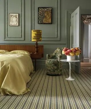 Grande chambre avec murs peints en vert et lambris, œuvres d'art encadrées et photos sur les murs, lit avec tête de lit en bois foncé, literie jaune, table ronde tulipe blanche, tapis à rayures, petit fauteuil avec revêtement à motifs floraux