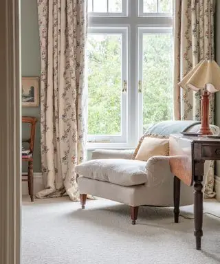 Chambre confortable avec tapis en laine crème, fauteuil décontracté avec coussins, table d'appoint en bois sombre avec lampe de table, grandes fenêtres blanches avec rideaux fleuris