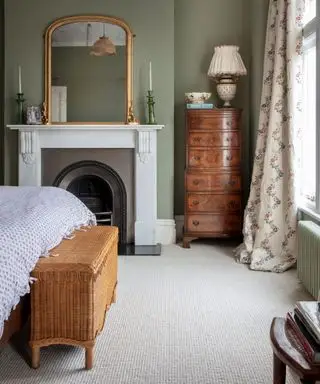 Chambre de style classique avec tapis crème, cheminée, murs peints en vert, armoire en bois sombre, rideaux crème fleuris, lit avec coffre de rangement en osier au pied