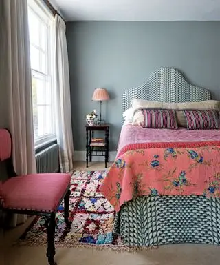 Chambre avec murs vert sauge, fenêtres à guillotine, literie lumineuse et tête de lit rembourrée, chaise rose, tapis à motifs sur moquette crème