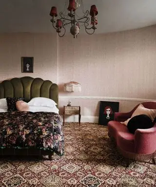 Chambre de style vintage avec moquette à motifs rouges, papier peint à motifs roses, plafond blanc, lustre avec abat-jour en tissu, lit avec tête de lit festonnée verte, canapé rouge foncé avec coussins