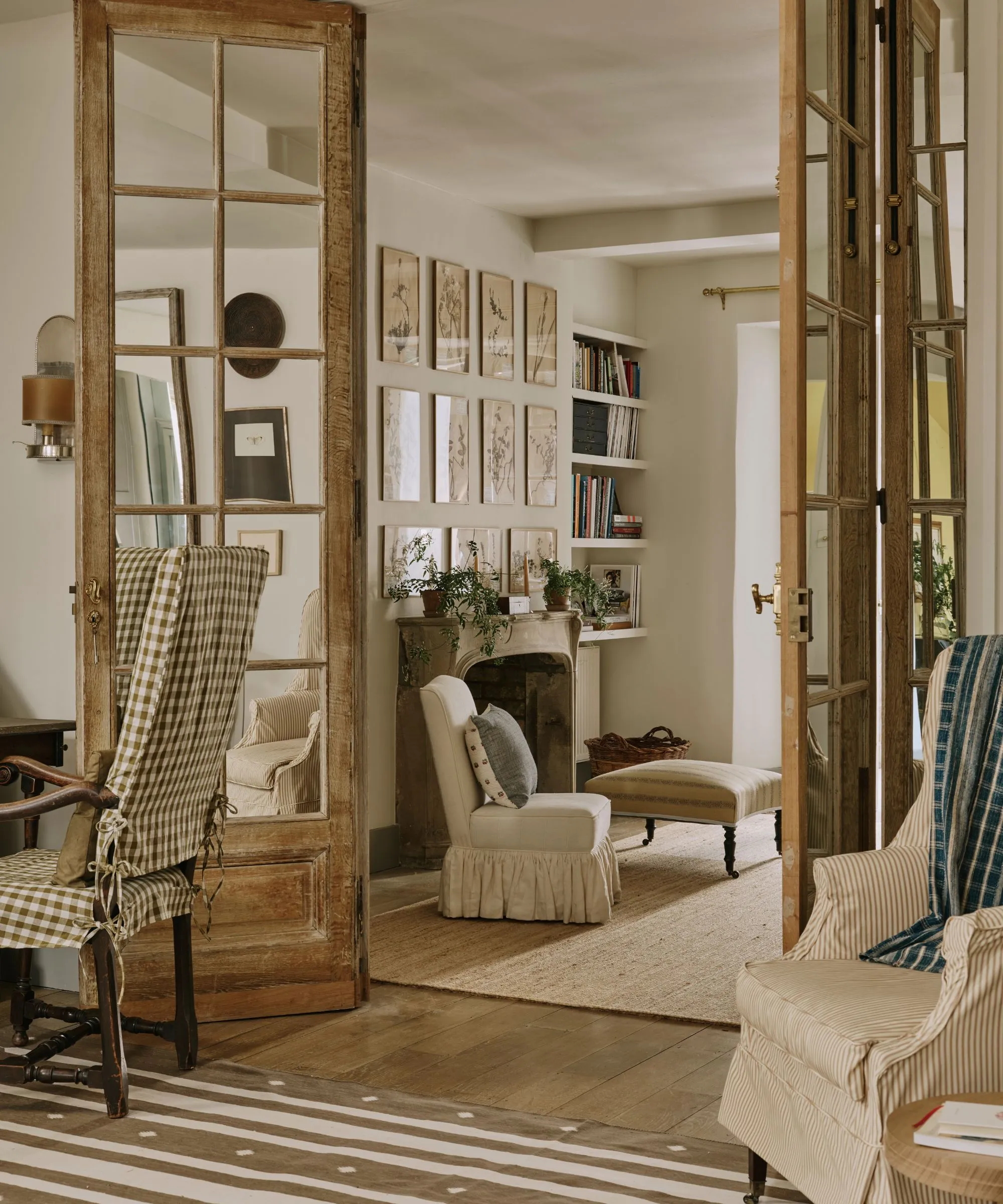 Pohľad cez zvetrané drevené francúzske dvere do pokojného, ​​neutrálne tónovaného obytného priestoru. Izba je vybavená kamenným krbom, stenou galérie botanických náčrtov a útulným potiahnutým nábytkom vrátane kresla s plisovanou sukňou a zeleno-bielej ginghamovej stoličky v popredí.' loading='lazy' title=