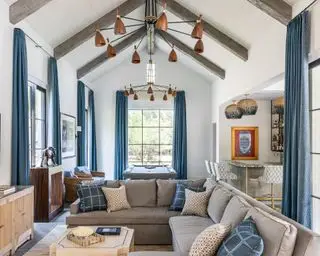 Grand salon avec plafond voûté, poutres en bois sombre et plafond lambrissé blanc, rideaux bleus, canapé d'angle gris, table de billard, meuble en bois avec télévision montée au-dessus, deux lustres en cuivre