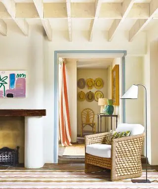 Maison de campagne moderne avec poutres à caissons, murs peints en crème, fauteuil avec lampadaire à côté, tapis à motifs, cheminée avec œuvres d'art modernes au-dessus, donnant sur le couloir avec table d'appoint, présentoir de chapeaux décoratif sur les murs, chaise en bois