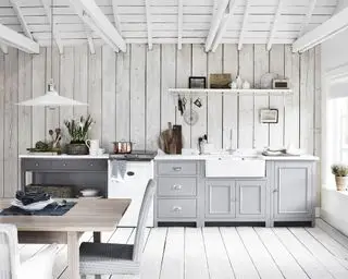 Espaces de cuisine blancs avec plafond en bois blanc, murs lambrissés blanc cassé, parquet en bois blanc, éléments de cuisine gris, suspension blanche, table à manger en bois avec chaises rembourrées