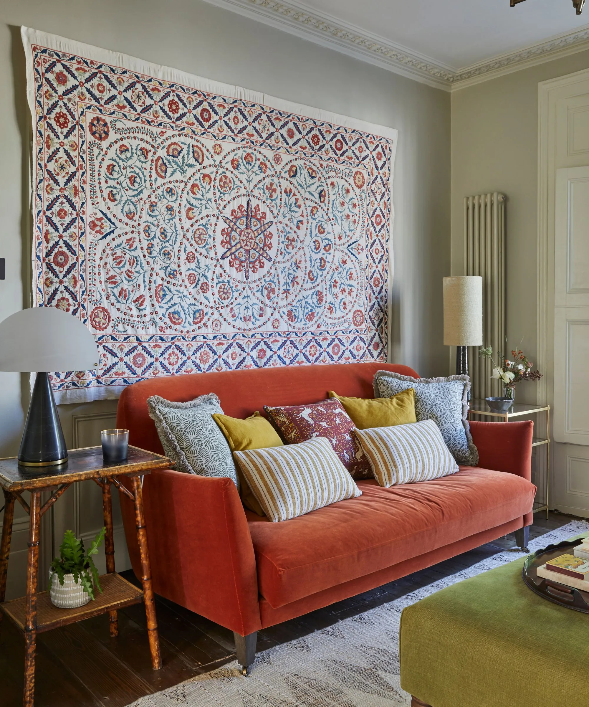 obývacia izba s červeným gaučom s množstvom vankúšov na vrchu, veľký gobelín visiaci zo steny za ním a bočnými stolíkmi po oboch stranách gauča' loading='lazy' title=