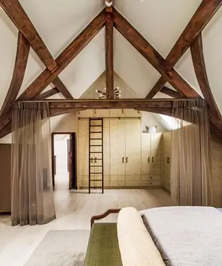 Chambre principale avec mur de rangement et échelle coulissante pour accéder au niveau supérieur, armoires et poutres apparentes sous un plafond voûté et voilages tirés.