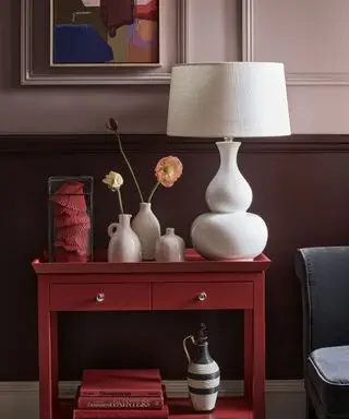 Chambre peinte en violet clair avec lambris peints en violet foncé, tableau peint sur le mur, abat-jour blanc, table d'appoint rouge avec livres et ornements décoratifs, chaise bleue
