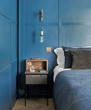 Chambre avec murs lambrissés bleus, lit double avec literie en denim bleu et coussins bleus et noirs, applique avec ampoule suspendue apparente, table de chevet en bois et parquet