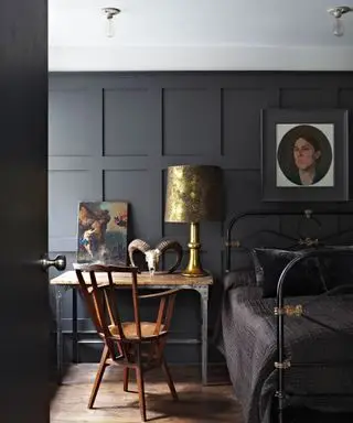 Chambre avec boiseries peintes en noir, cadre de lit en métal noir avec couette noire, bureau en bois et métal avec fauteuil traditionnel en bois foncé, peintures sur le bureau et sur les murs, abat-jour métallique