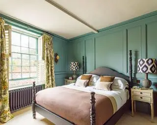 Chambre principale avec boiseries bleu-vert et lit double, rideaux fleuris jaunes et verts, tables de chevet doubles et lampes de table avec abat-jour à imprimé géométrique, cadre de lit en bois noir, literie et coussins blancs et rose pâle, moquette crème