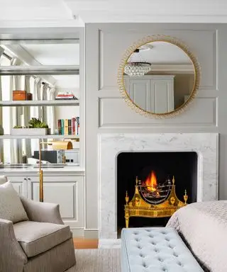 Chambre décorée en gris pâle avec un feu allumé dans une cheminée en marbre, un tapis gris pâle, un fauteuil et un pouf bleu.