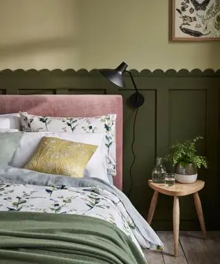 Chambre avec lambris vert foncé à bordure festonnée, murs peints en vert clair, applique noire, tabouret en bois clair, literie à fleurs vertes et grises avec tête de lit rose pâle, coussins dispersés