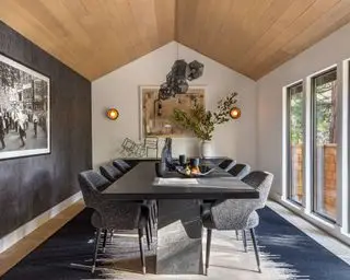 Salle à manger avec plafond voûté, revêtement en bois chaleureux, grande table à manger en marbre noir avec chaises rembourrées, luminaire suspendu moderne, tapis bleu sur parquet, mur texturé gris avec œuvres d'art
