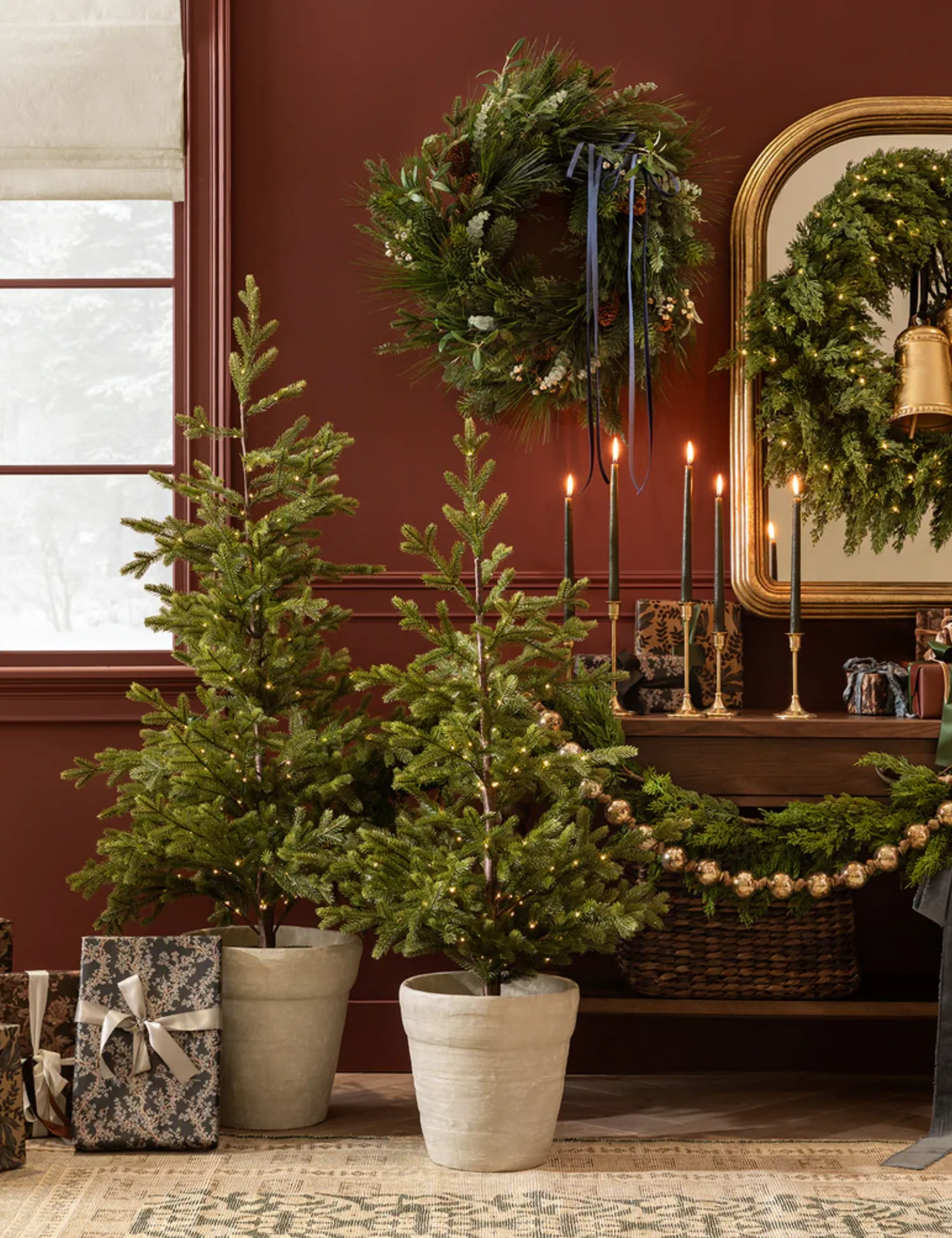 Des mini sapins de Noël Shea McGee décorés dans un salon bordeaux aux côtés de couronnes de Noël, de chandeliers dorés et de cadeaux emballés.' loading='lazy' title=