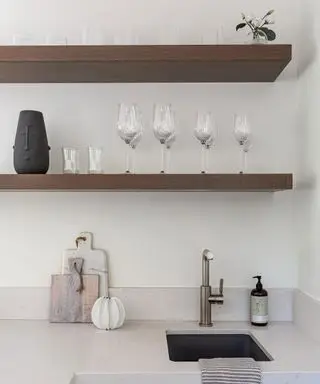 キッチンシンク上に配置された2つの木製棚を中心としたニュートラルなキッチンスペース。ガラス製品、装飾的な花瓶、植物を使ったミニマルなスタイル。天然のクリームストーンのカウンタートップ、大理石と木製のまな板、ハンドソープ、銀の蛇口