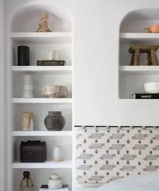 壁に埋め込まれた 2 つのアーチ型の棚、白く塗られたテクスチャーのある壁、本が飾られた棚、装飾品の花瓶など、すべてが落ち着いたニュートラルなカラーパレットでまとめられています。