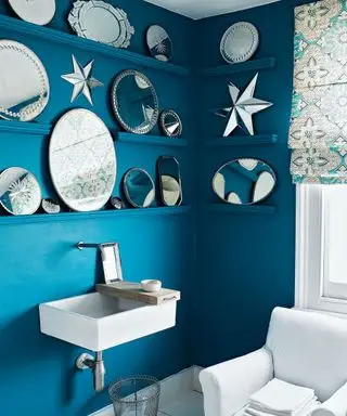 青く塗られたバスルームには、棚に華やかな鏡を選択でき、長方形のシンク、木の板、白いアンティークの肘掛け椅子、タオル、ワイヤーくずかご、白と青の模様のブラインドが備わっています。