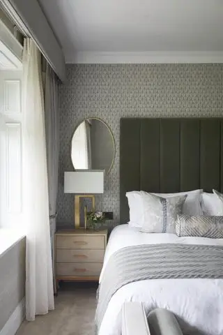 chambre grise avec papier peint à motifs gris, tête de lit en velours vert sauge, linge de lit blanc, miroir doré, lampe de table dorée, rideaux gris