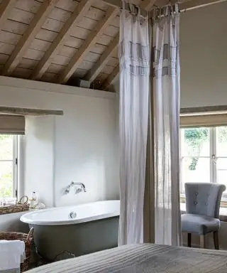 Chambre rustique et campagnarde avec poutres apparentes sur plafond voûté en bois, lit à baldaquin en bois avec rideaux transparents doux, baignoire à côté de la fenêtre