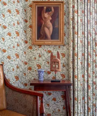 Gros plan sur un papier peint orange et vert et des rideaux assortis, une photo encadrée en or, une table d'appoint et un fauteuil en bois foncé