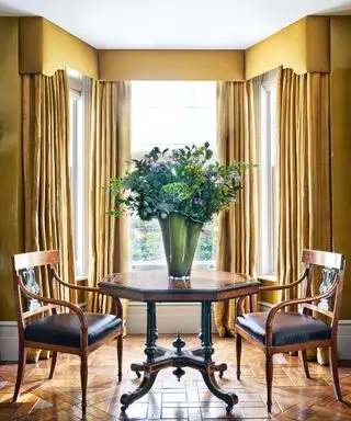Salon classique avec table et chaises en bois sombre, murs peints en moutarde et rideaux assortis avec cantonnière sur une grande fenêtre, grand vase avec fleurs sur la table
