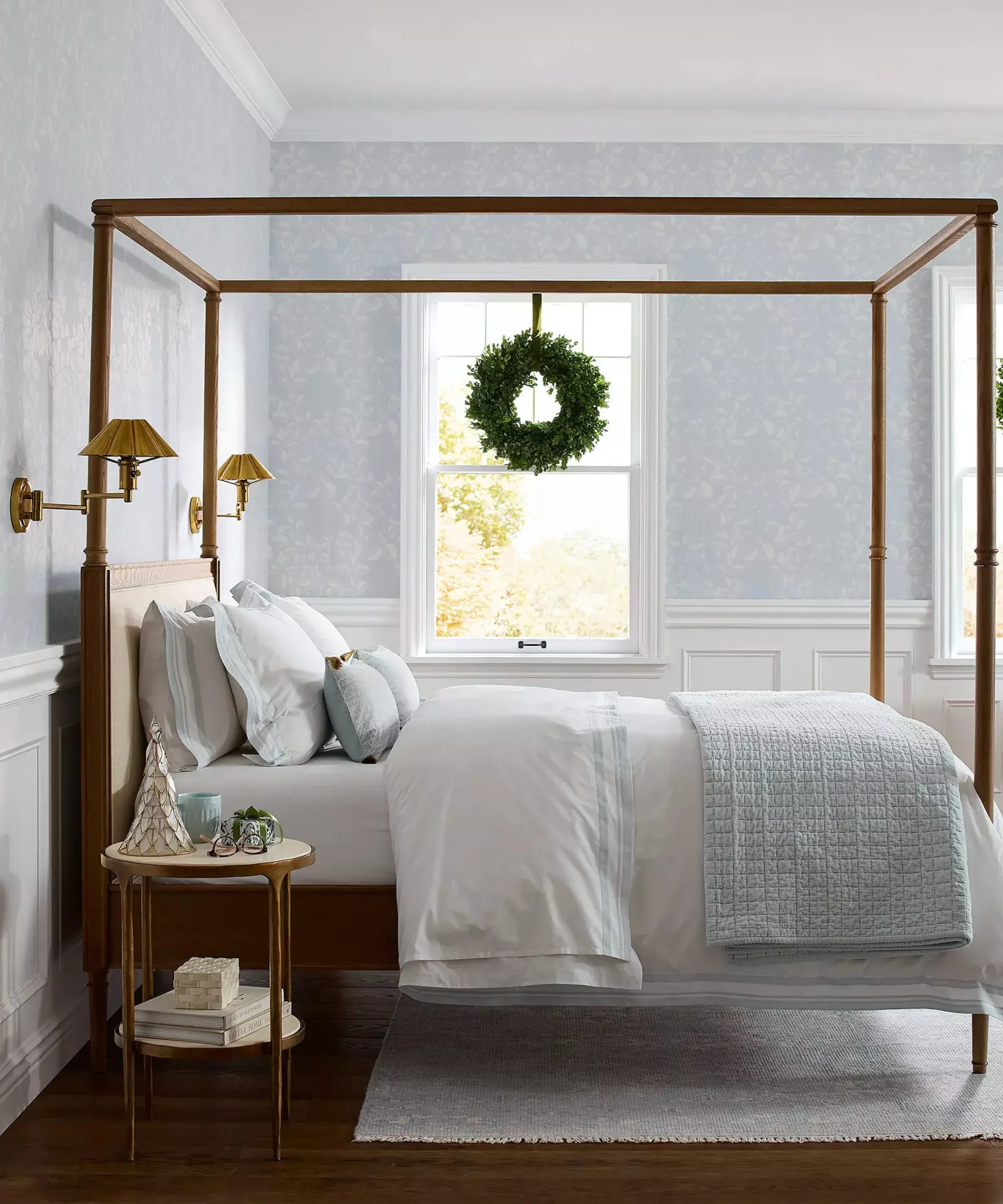 nadmorska sypialnia z niebieską tapetą, łóżko z ciemną drewnianą ramą, jasnoniebieska i biała pościel oraz wieniec zwisający z okna' loading='lazy' title=