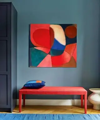 나무 바닥과 파란색 깔개로 마감된 파란색 페인트 복도. 밝은 빨간색 벤치, 벤치 위 벽에 장착된 대담하고 다채로운 예술 작품, 구석에 진한 파란색으로 칠해진 캐비닛