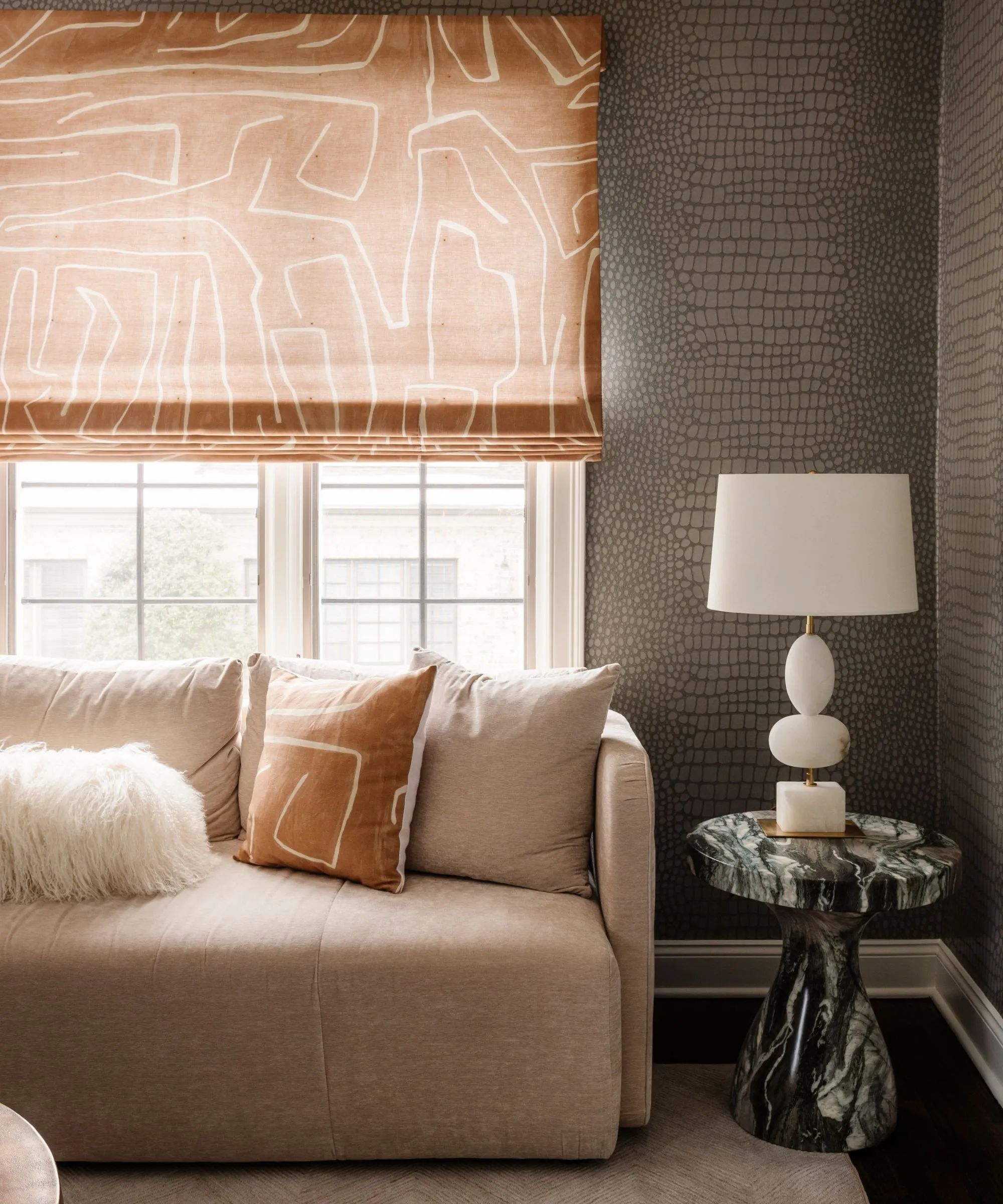 壁にヘビ皮の壁紙が貼られたリビング ルーム、ベージュのソファ大理石のサイド テーブルと白いランプ、ソファの上の窓に模様の入ったオレンジ色のブラインド' loading='lazy' title=