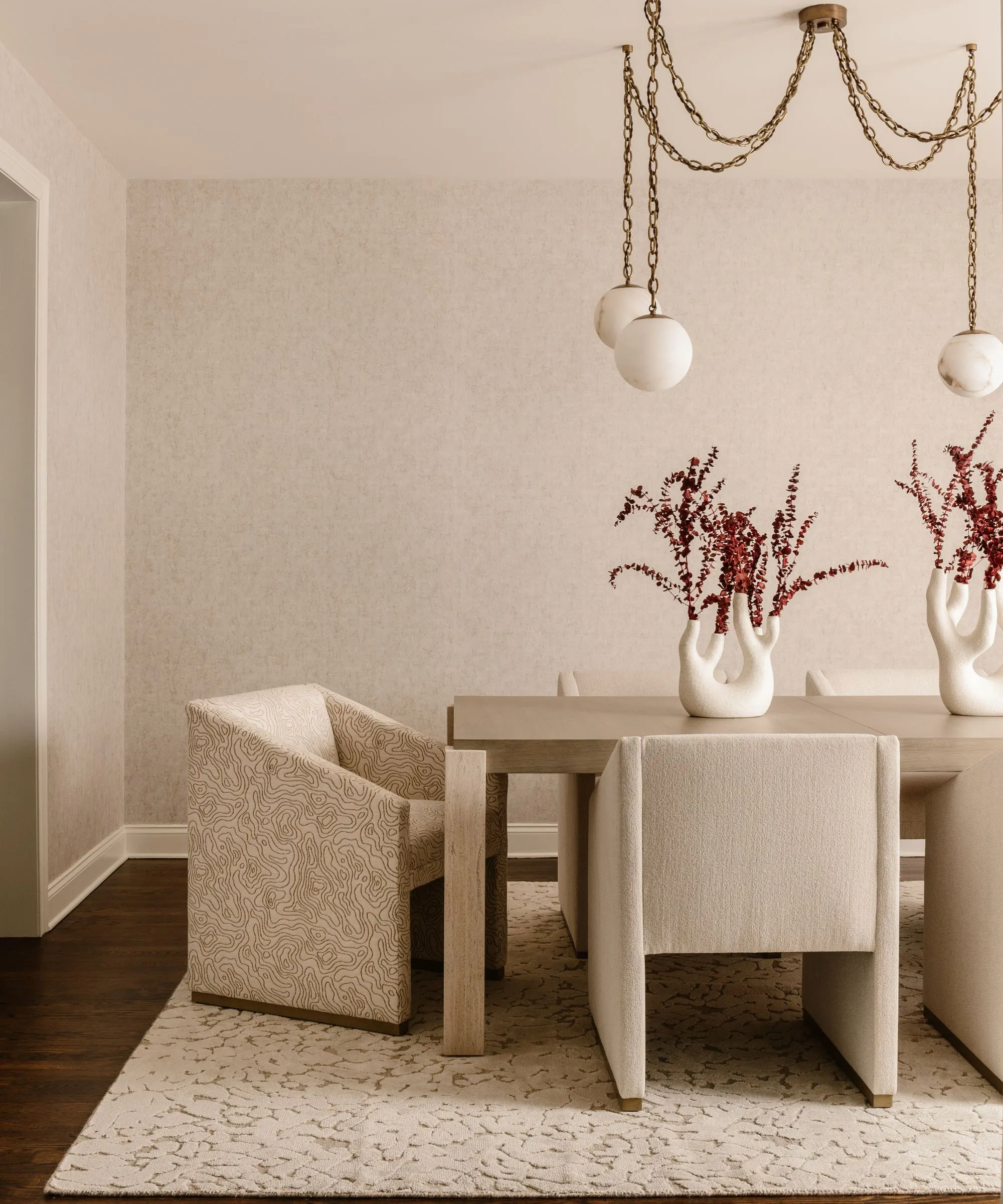 ベージュのダイニング ルームには、ベージュとベージュの模様の木製テーブルと布張りのダイニング チェアが置かれています。テクスチャーのベージュの敷物がダイニング テーブルの下にあり、ステートメントのゴールドのライトが上にぶら下がっています。' loading='lazy' title=