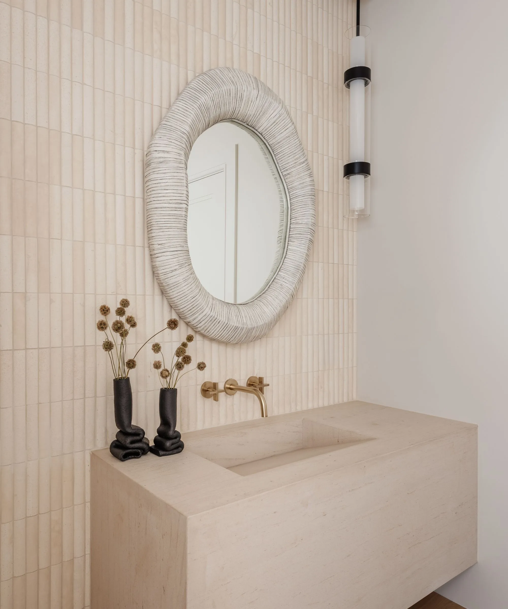 ニュートラルなタイルと石造りのシンクを備えたパウダールーム、その上にテクスチャーのある鏡があり、片側に黒いペンダント照明器具が吊り下げられています' loading='lazy' title=